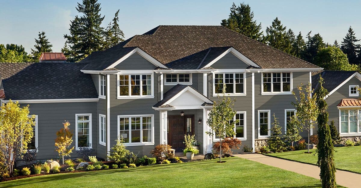 A large, modern gray house with white trim and a dark roof is surrounded by a well-maintained lawn and trees, exuding a peaceful and inviting vibe.