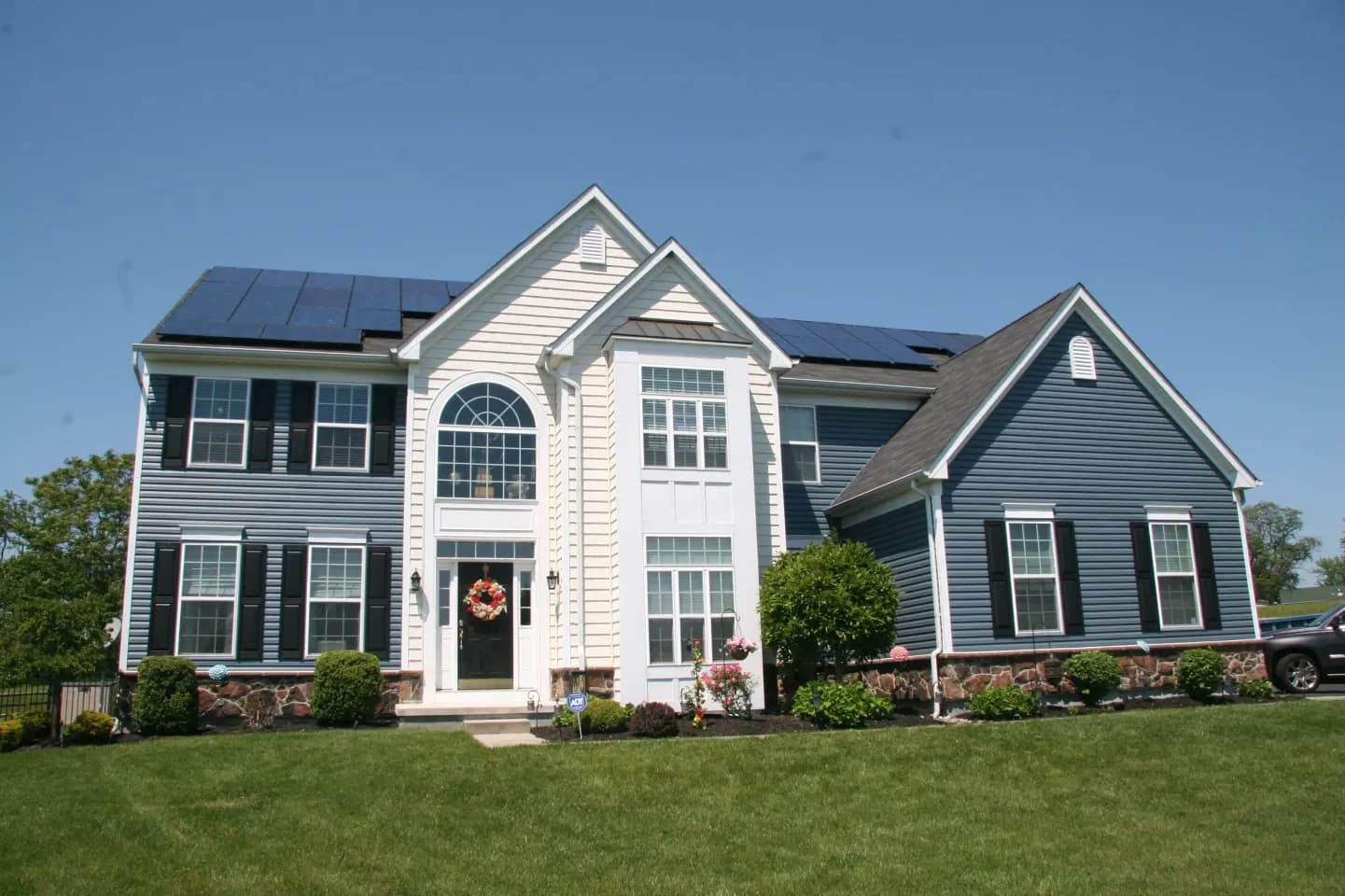 Two-story house with blue siding and white trim. Solar panels on the roof. Well-maintained lawn, shrubbery, and a wreath on the front door. Bright sky.
