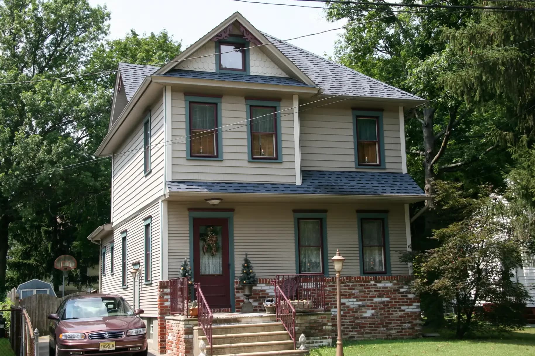 Two-story beige house with red trim, surrounded by lush trees. A car is parked in the driveway. The front features a small porch with steps and festive wreaths, creating a cozy atmosphere.
