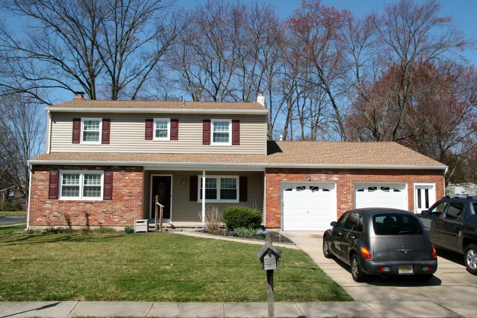 Two-story suburban house with brick and beige siding, a double garage, and a driveway with two cars. Leafless trees and a blue sky in the background.